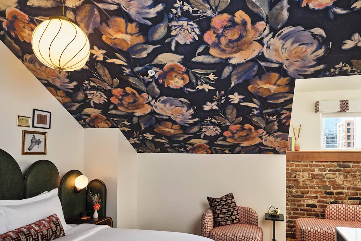 Une chambre d’hôtel avec un grand lit, un coin salon avec des fauteuils à motifs, une petite table et un mur en briques.