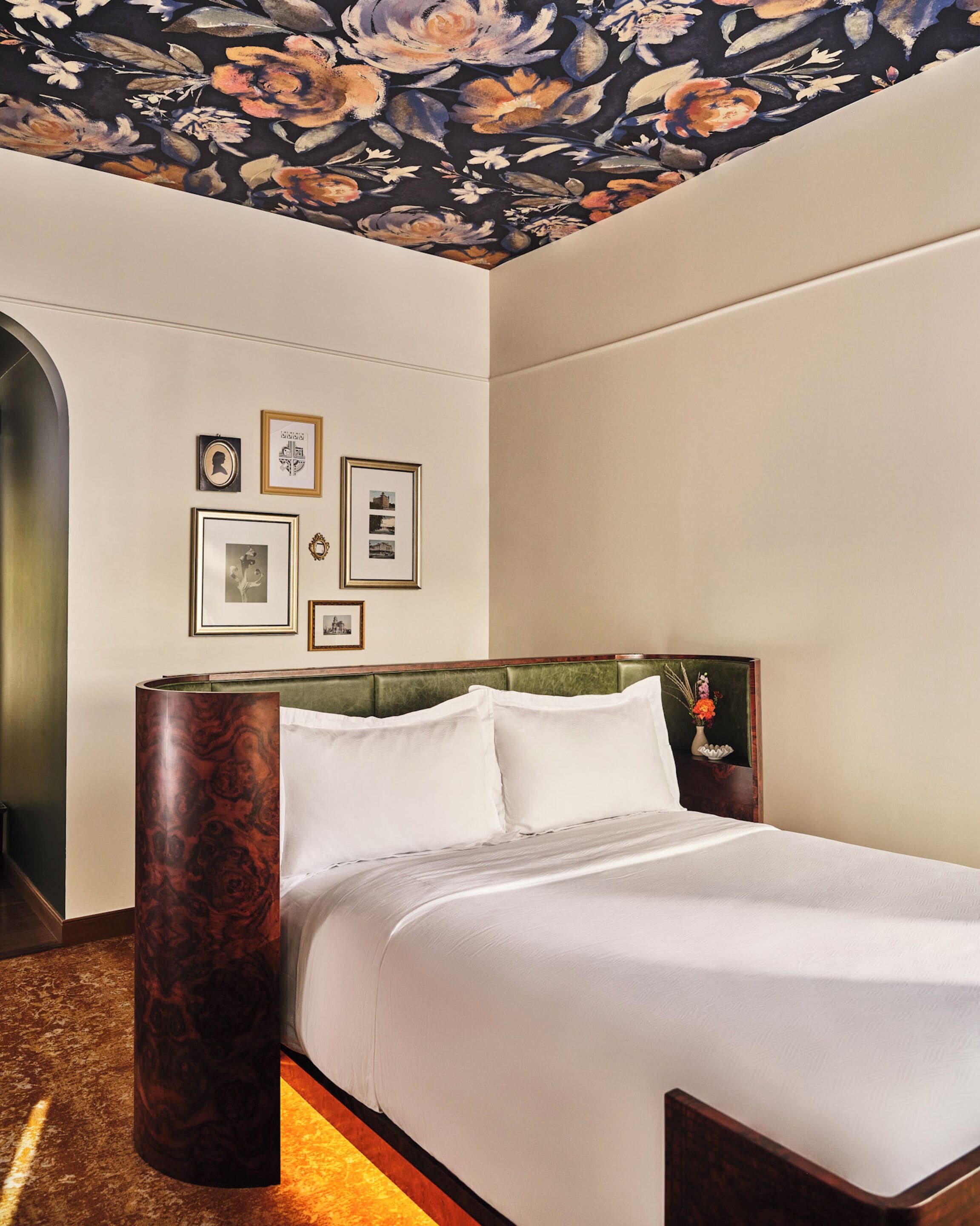 Habitació d'hotel amb un llit gran, un panell de capçalera tapissat de verd i un sostre amb motius florals. Hi ha quadres amb marc a la paret i un alt pilaret de fusta fosca.