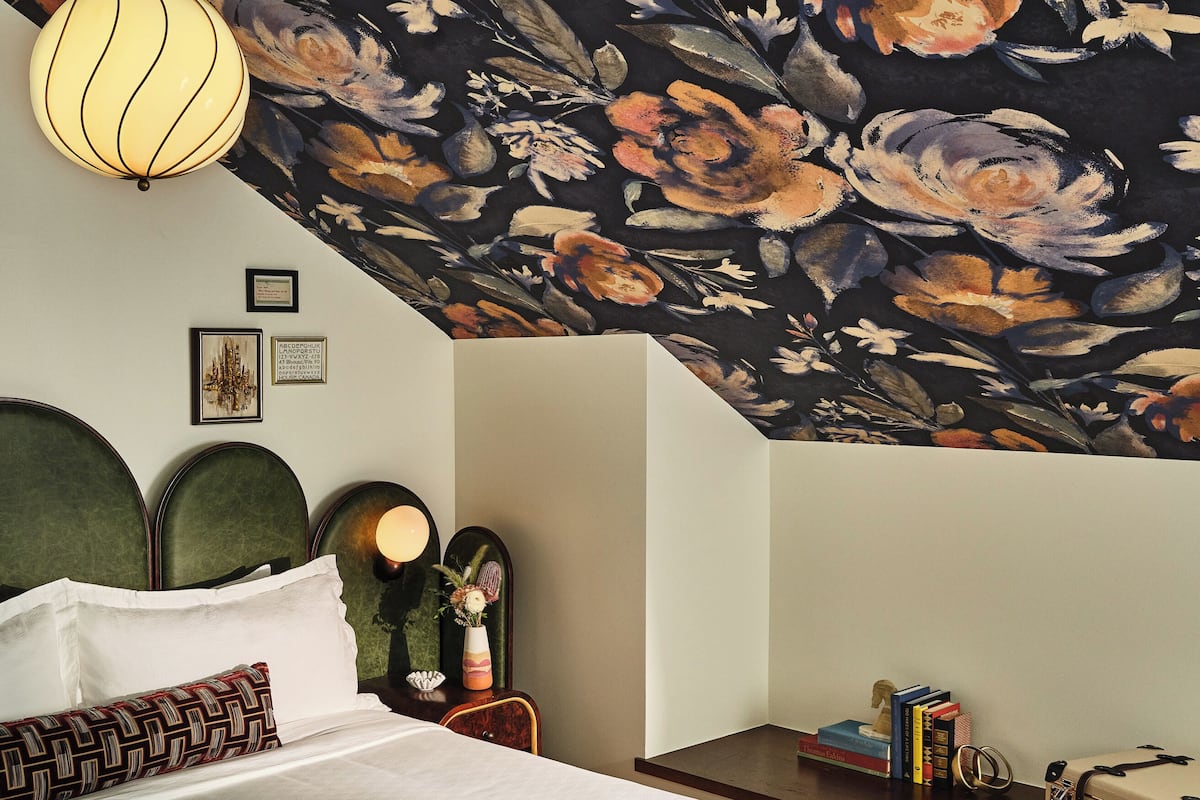 Une chambre à coucher avec un grand lit, un banc, une table de chevet avec des livres et un plafond recouvert d’un papier peint à motifs floraux.