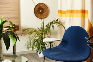 Una habitación con una silla azul, una mesita blanca pequeña, una planta en maceta y un plato decorativo colgado en la pared.