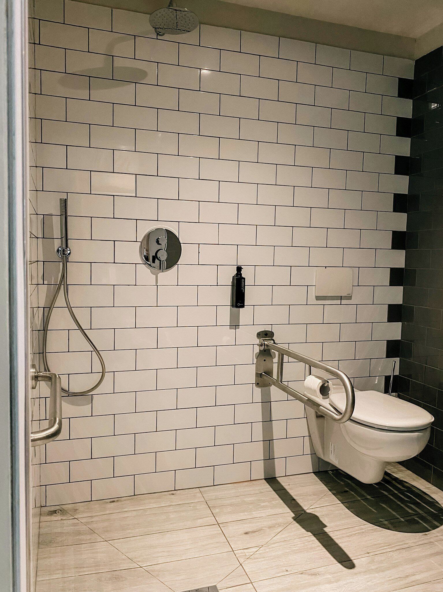 Baño con paredes de azulejos blancos, ducha, inodoro y barandilla.