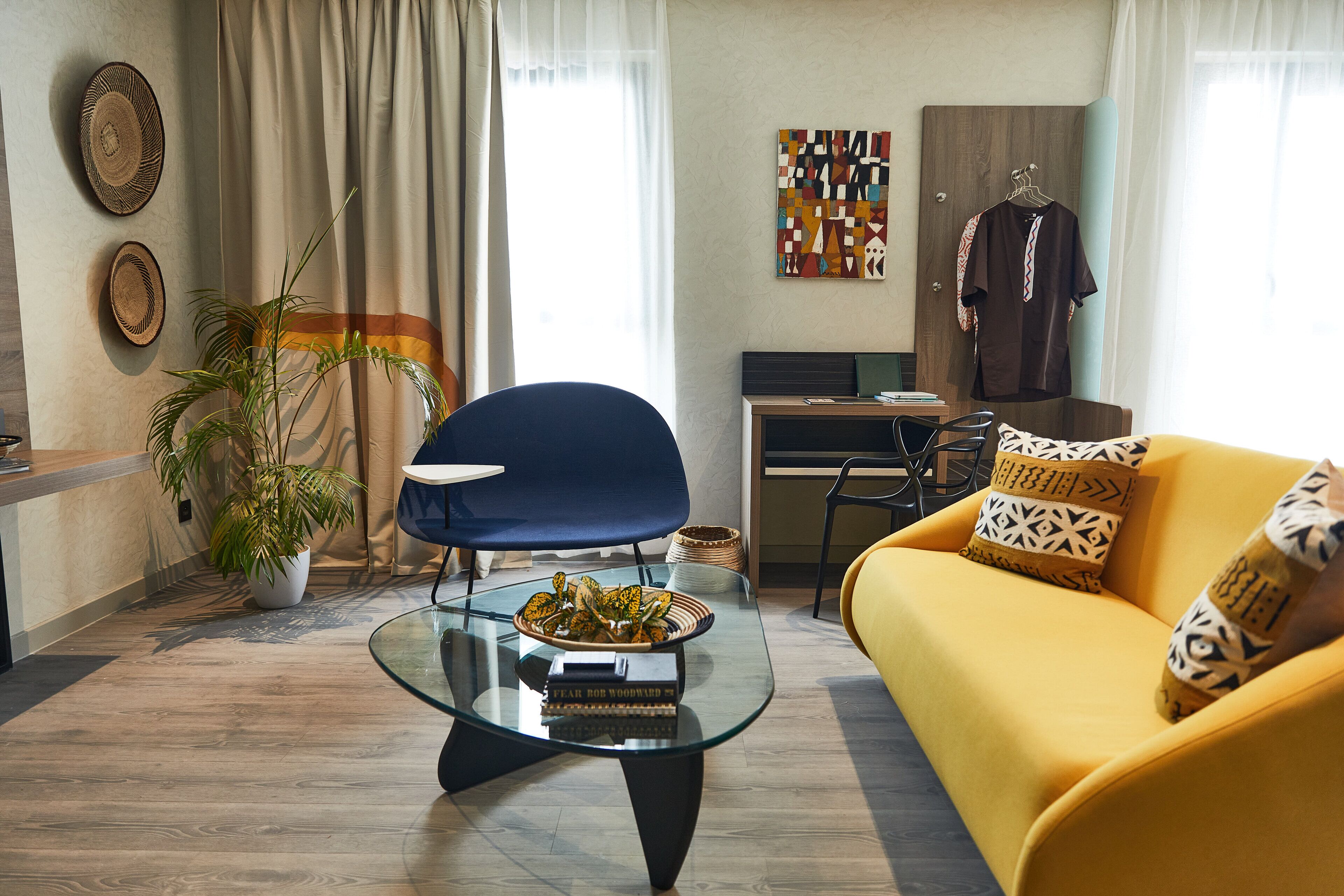 A modern living room with a glass coffee table, a yellow sofa, a blue chair, and a desk with a computer.