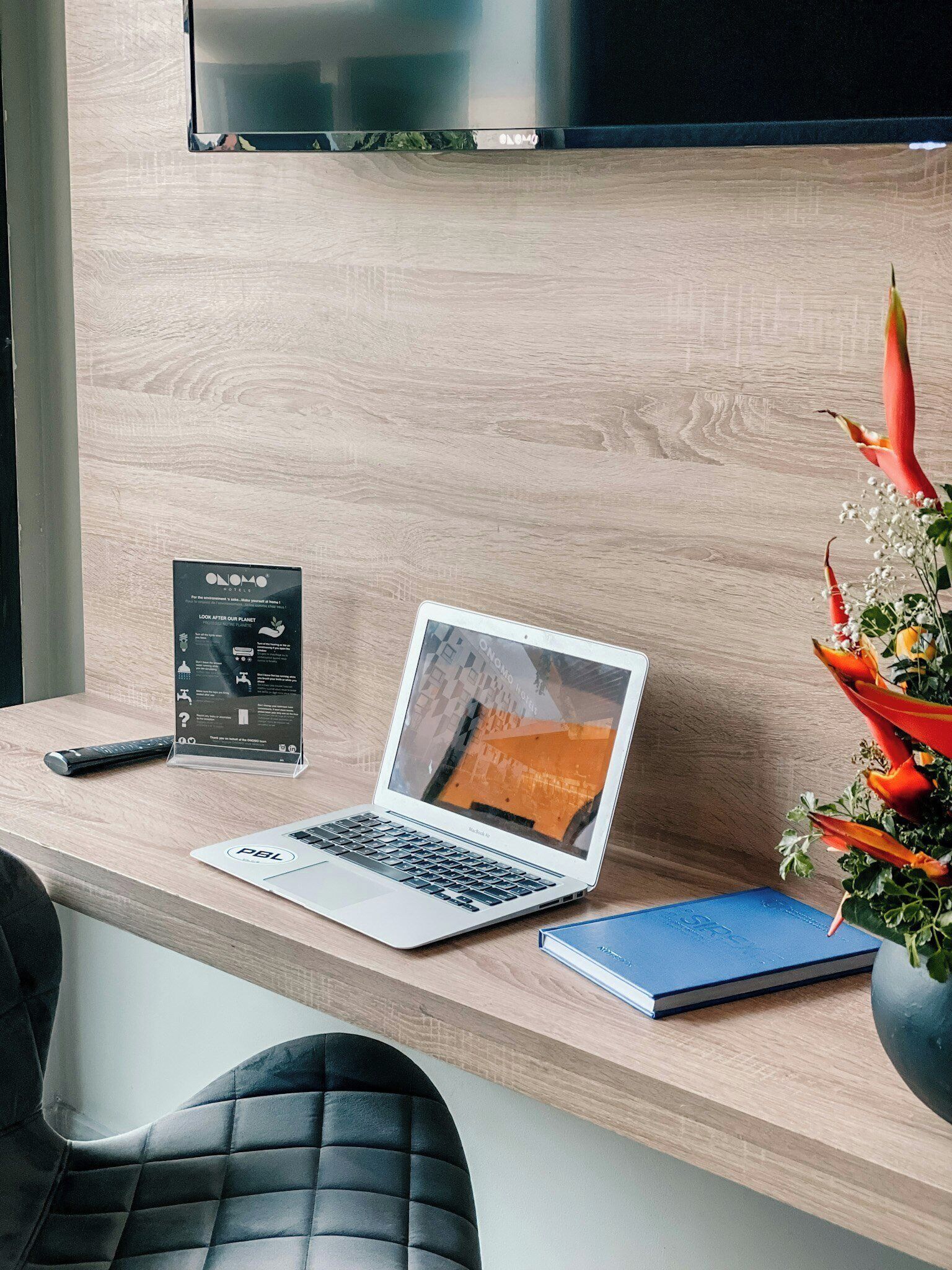 A laptop on a wooden desk with a remote control, a brochure, and a vase with flowers.