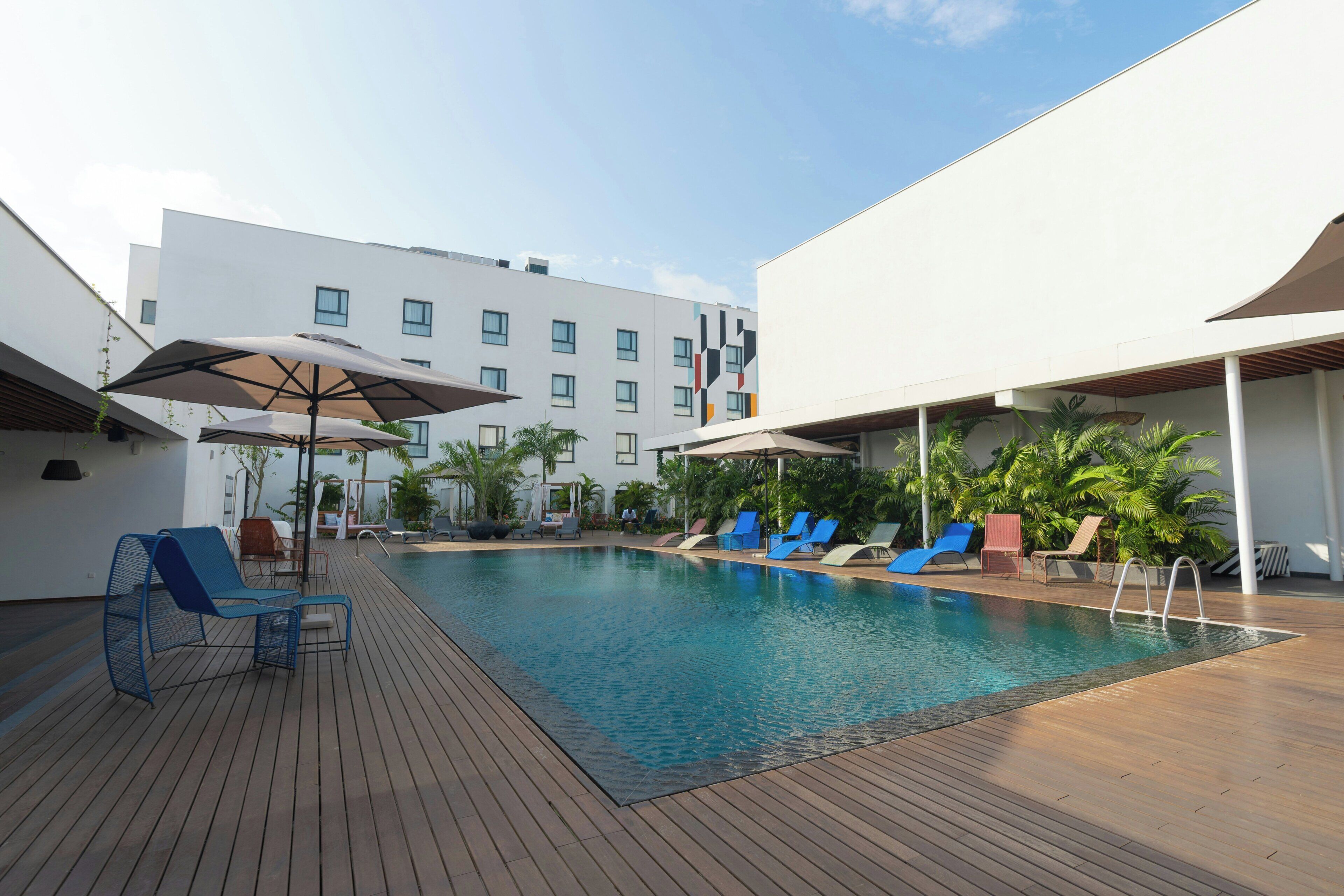 Une piscine entourée d’une terrasse en bois et d’un bâtiment doté de nombreuses fenêtres, avec des chaises longues et des parasols à proximité.