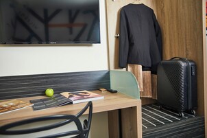 Habitación de hotel con escritorio, una silla, maleta negra, abrigo colgado y televisor en la pared.