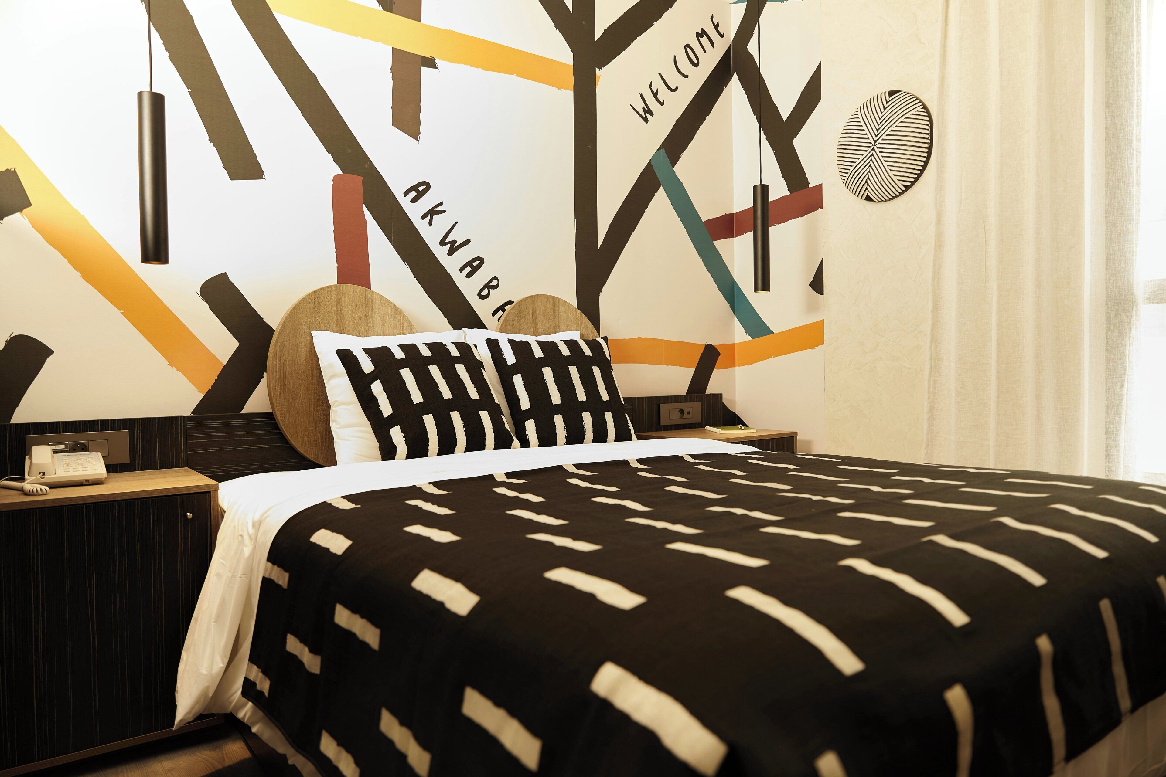 Une chambre d’hôtel avec un lit, une table de chevet avec un téléphone et un mur décoré d’œuvres d’art abstraites et du mot «BIENVENUE».