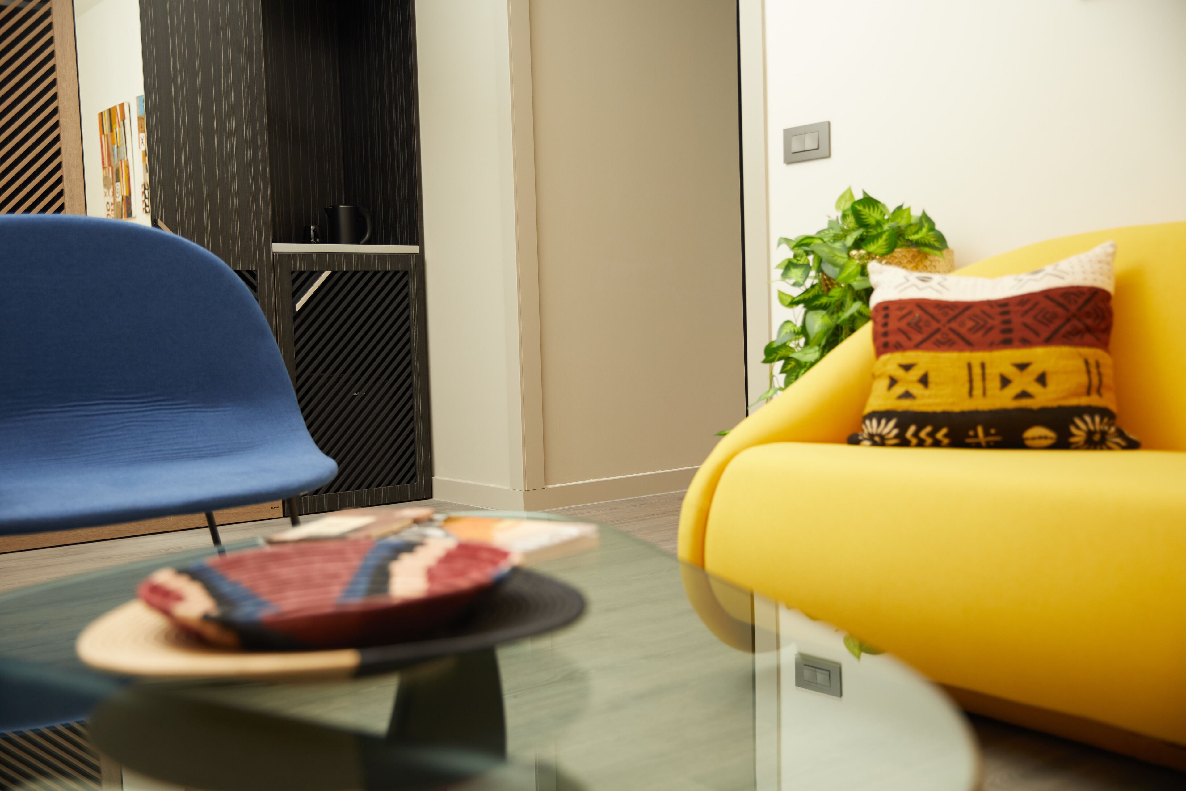 A modern living room with a yellow sofa, a blue chair, and a glass coffee table with a book and a plate.