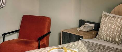 Una habitación de hotel con una cama, un sillón rojo, una mesita de noche con un teléfono y una colcha estampada.