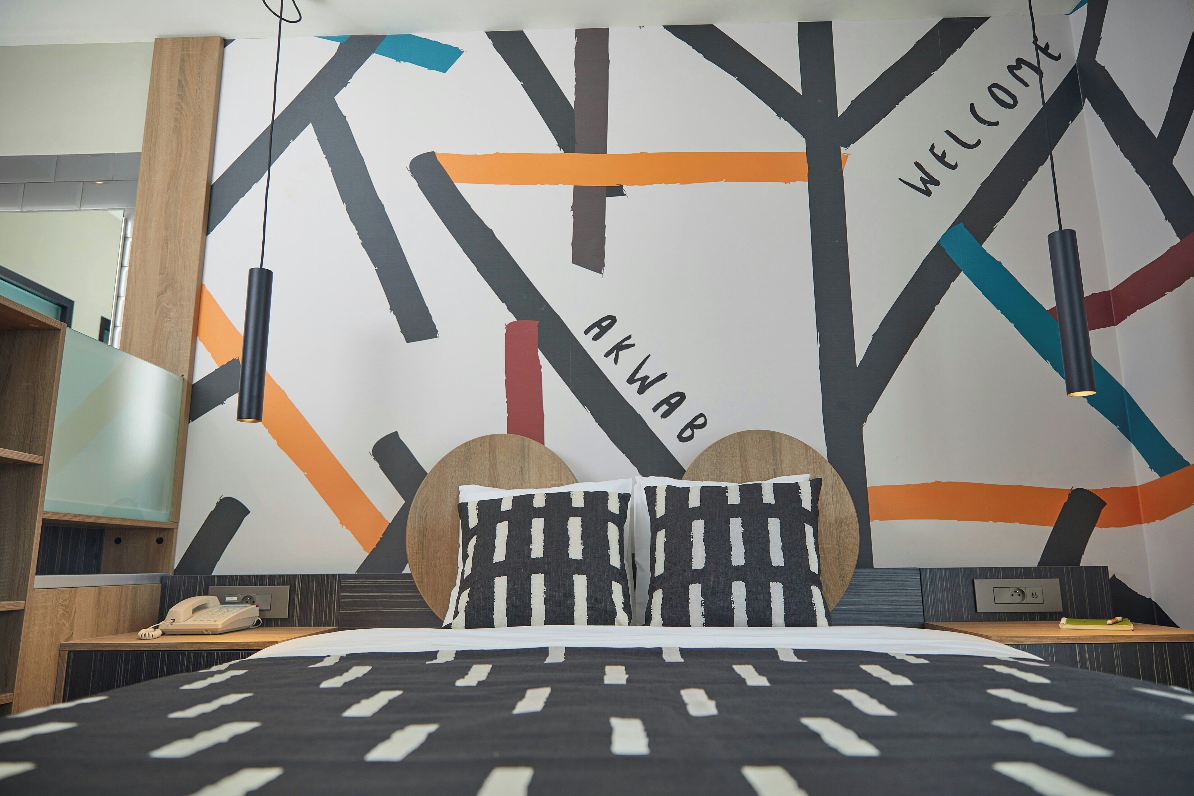 Een moderne hotelkamer met een bed, nachtkastje, telefoon en een muur met abstracte kunst en de woorden 'Welcome' en 'Akwaab'.