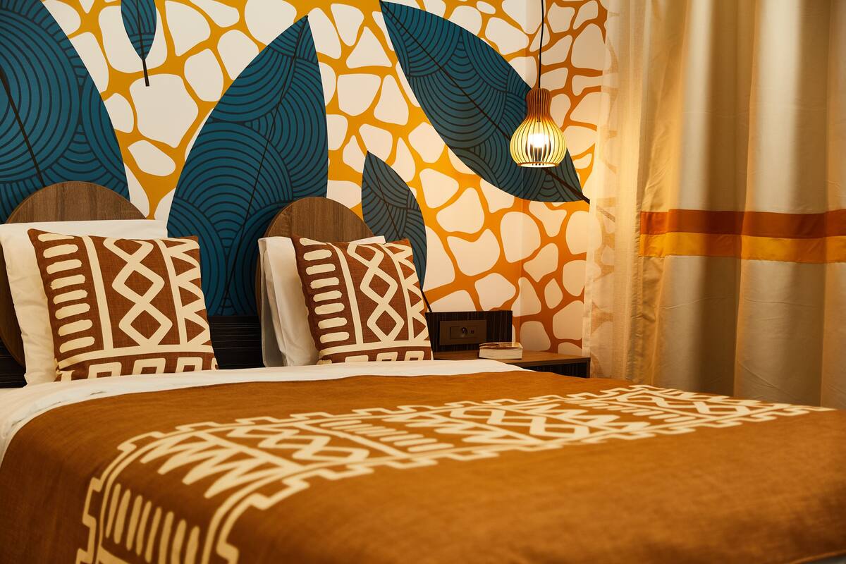 Een bed met beddengoed met een patroon, een nachtkastje met een lamp en een muur met een bladmotief.