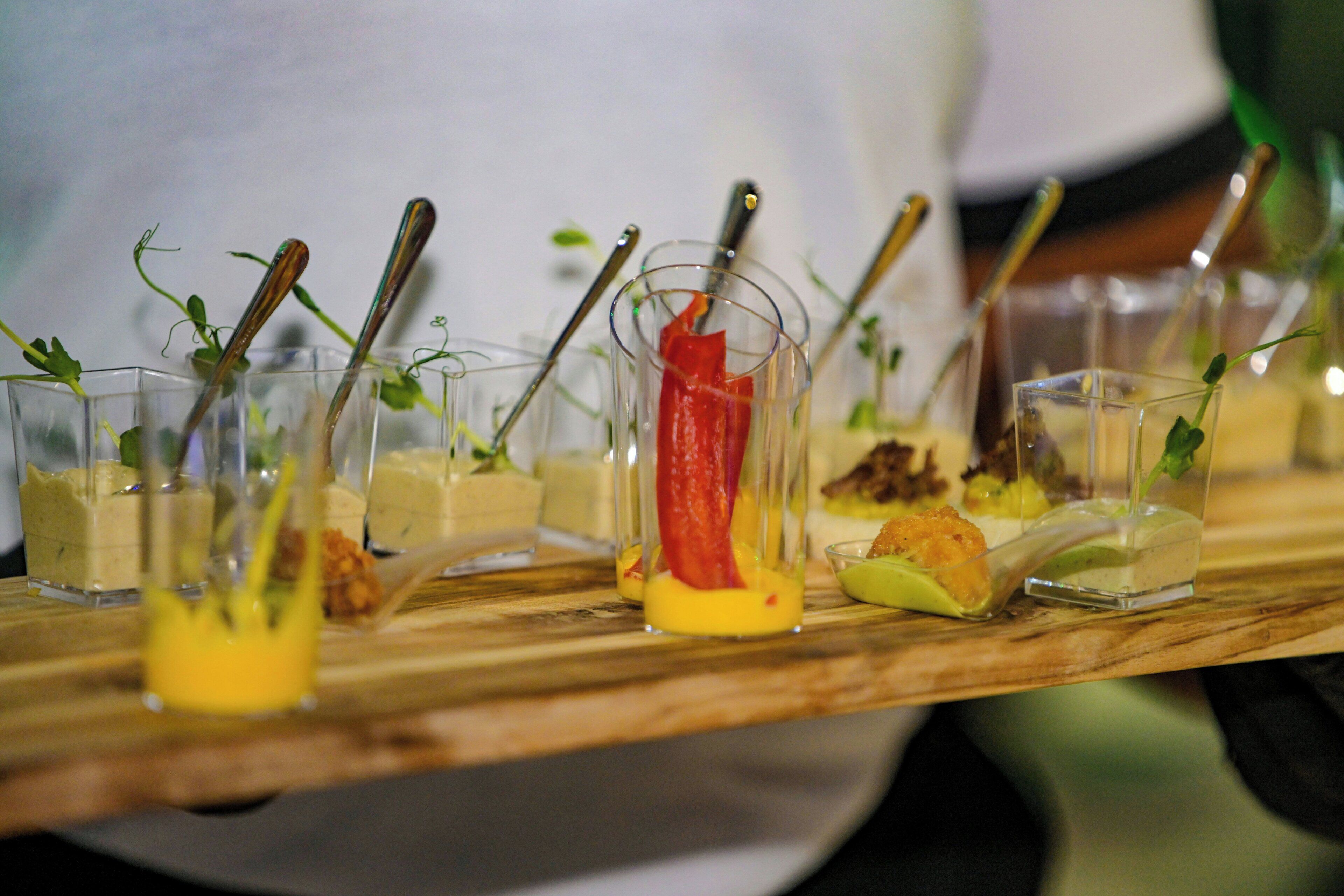 A wooden serving board with various food items in clear glasses, each with a spoon.