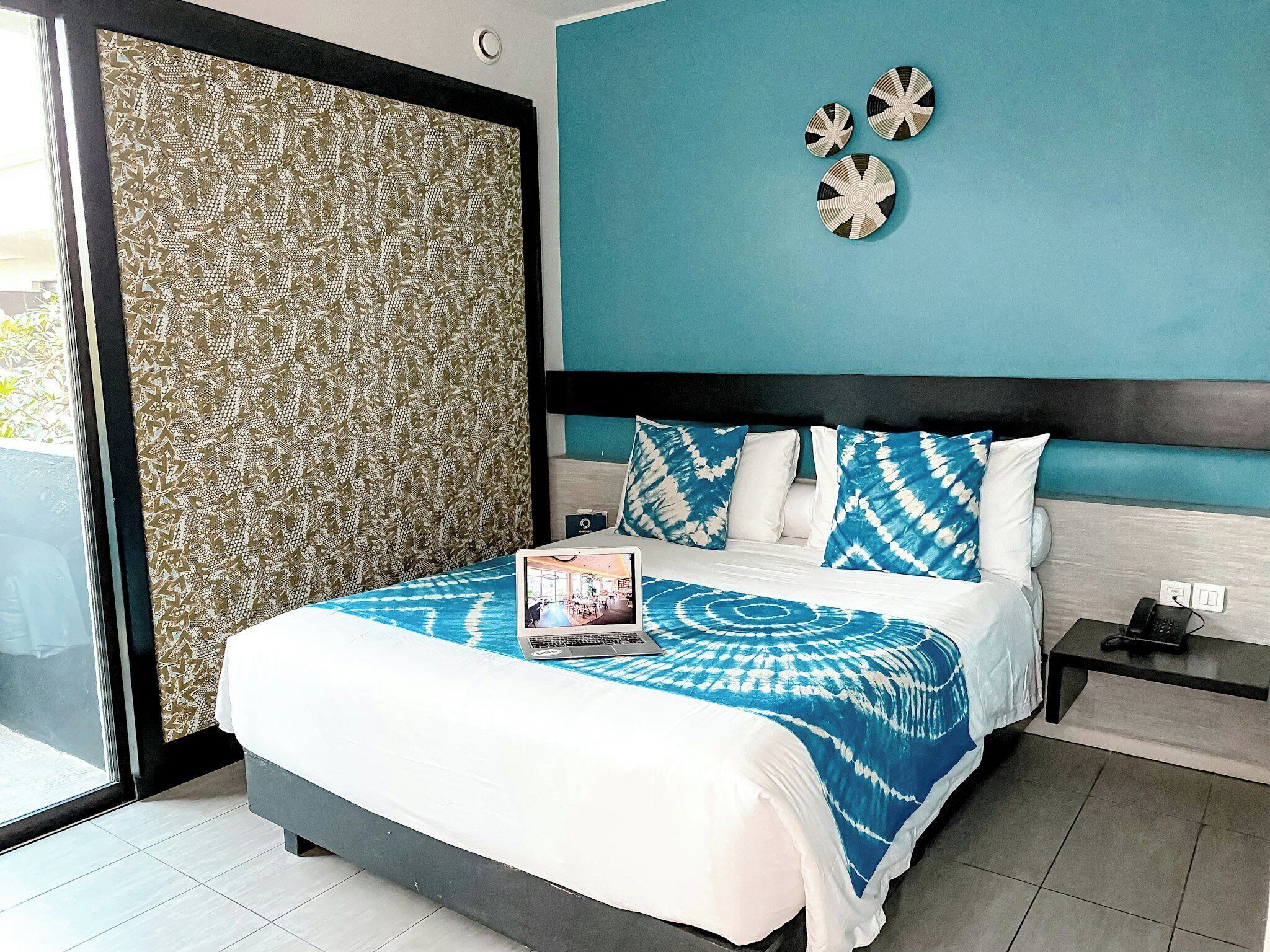 A modern hotel room with a bed, a laptop, a nightstand, and a view through a large window.
