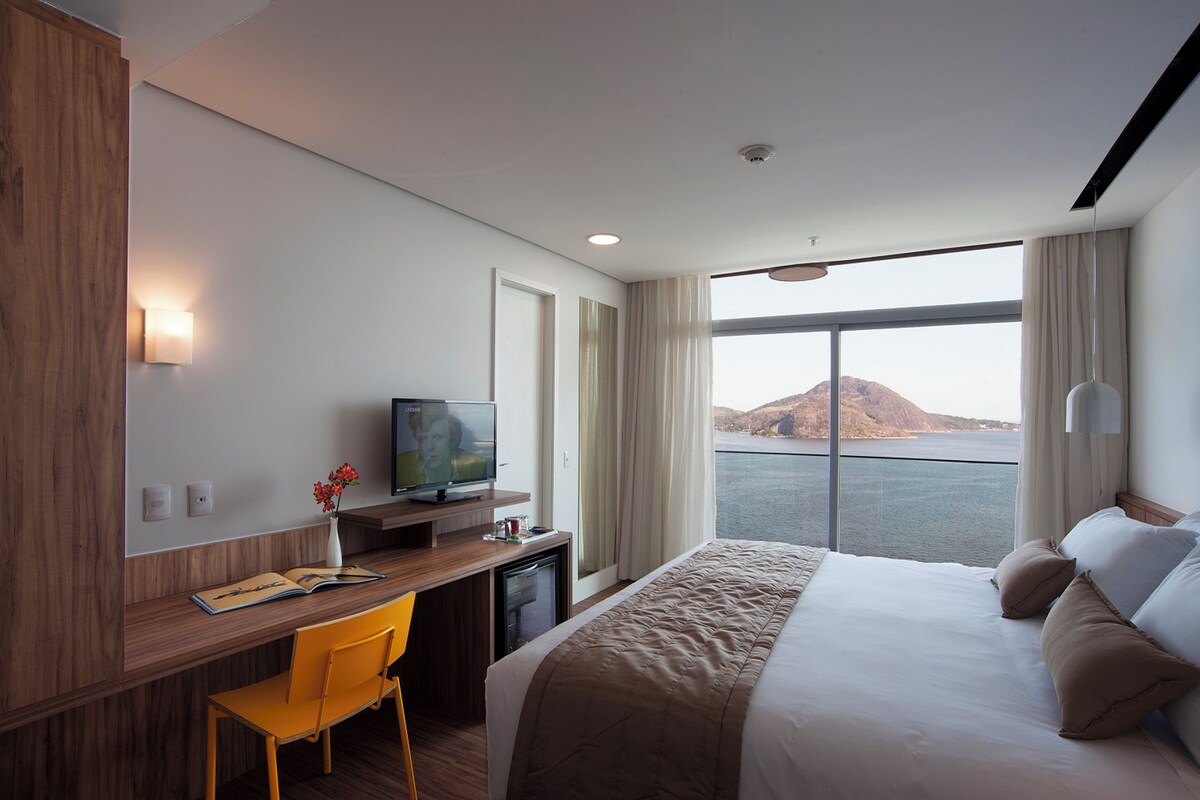 Une chambre d’hôtel moderne dotée d’un grand lit, d’un bureau avec une chaise, d’une télévision et offrant une vue sur une étendue d’eau, avec une montagne en arrière-plan.