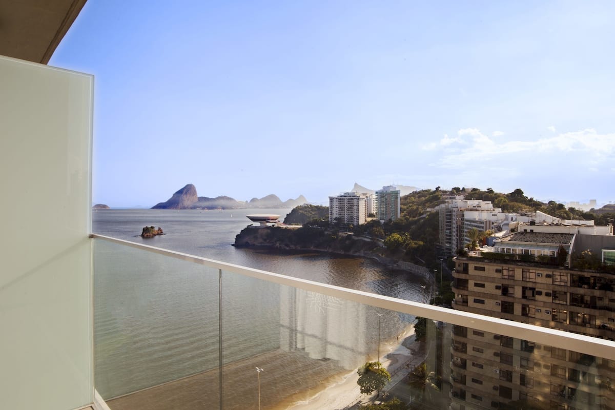 A view from a balcony overlooking a coastal cityscape with buildings, a beach, and a large rock formation in the distance.