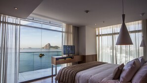 Un dormitorio moderno con una cama grande, un televisor sobre un mueble y vista al mar a través de ventanas de piso a techo.