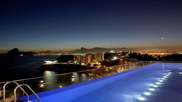 Una piscina en la azotea con vistas a un paisaje urbano al anochecer, con edificios iluminados y una montaña distante.