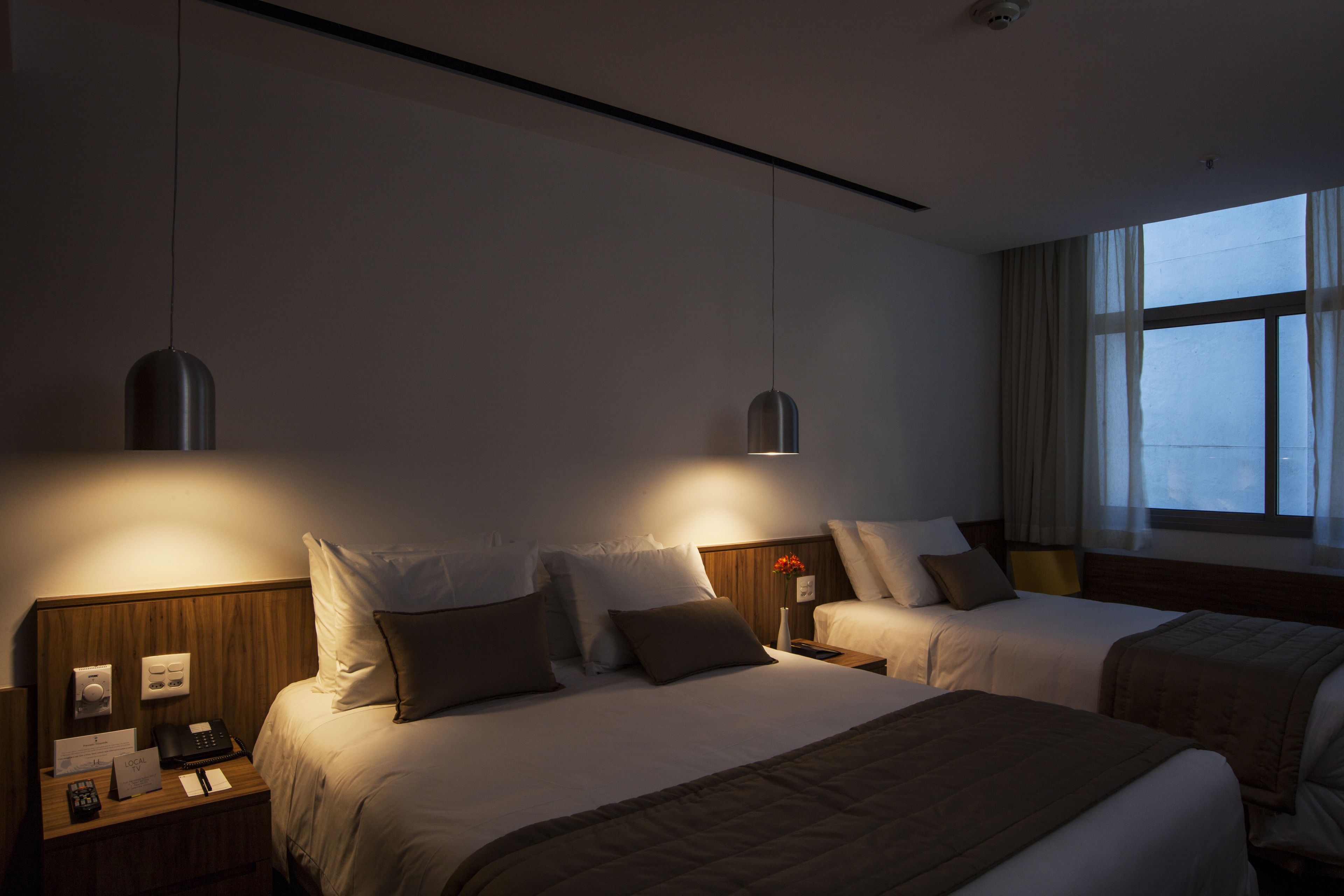 Habitación de hotel con dos camas, cabecero de madera, mesitas de noche con lámparas y una ventana con cortinas.