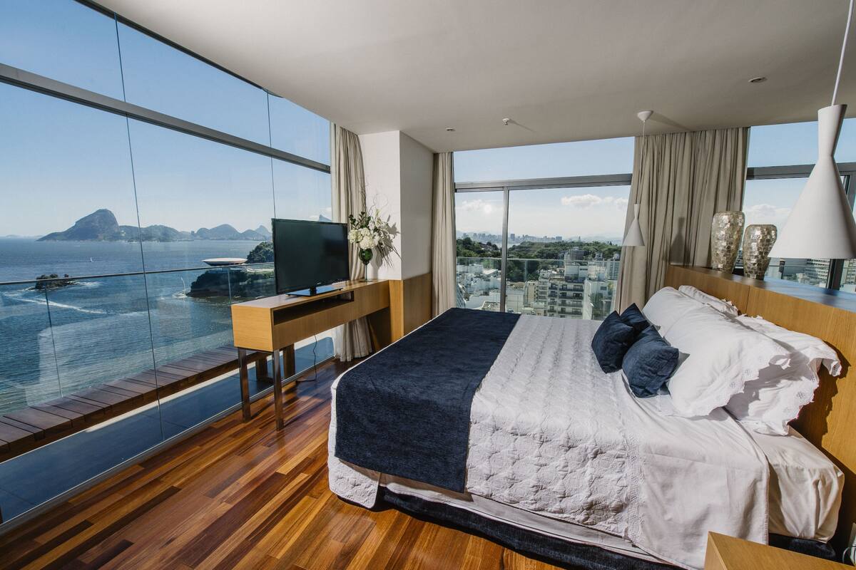 A modern bedroom with a large bed, a desk, and a TV. It features a balcony with a view of the sea and cityscape.