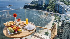 Una mesa con frutas, pan y una bebida, con vista a la playa y al paisaje urbano.