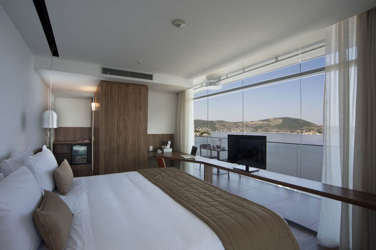 A hotel room with a large bed, a desk with a TV, and a balcony with a view of the sea.