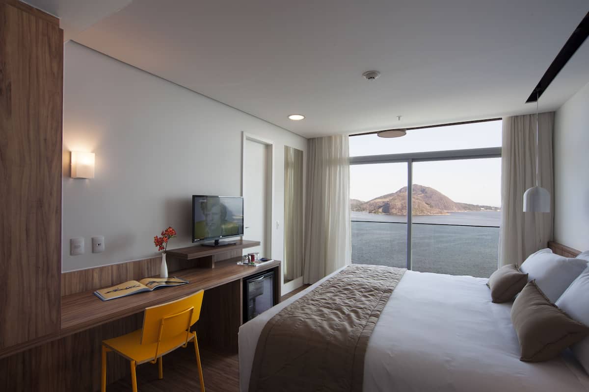 A hotel room with a bed, a desk with a chair, a television, and a view of the water.