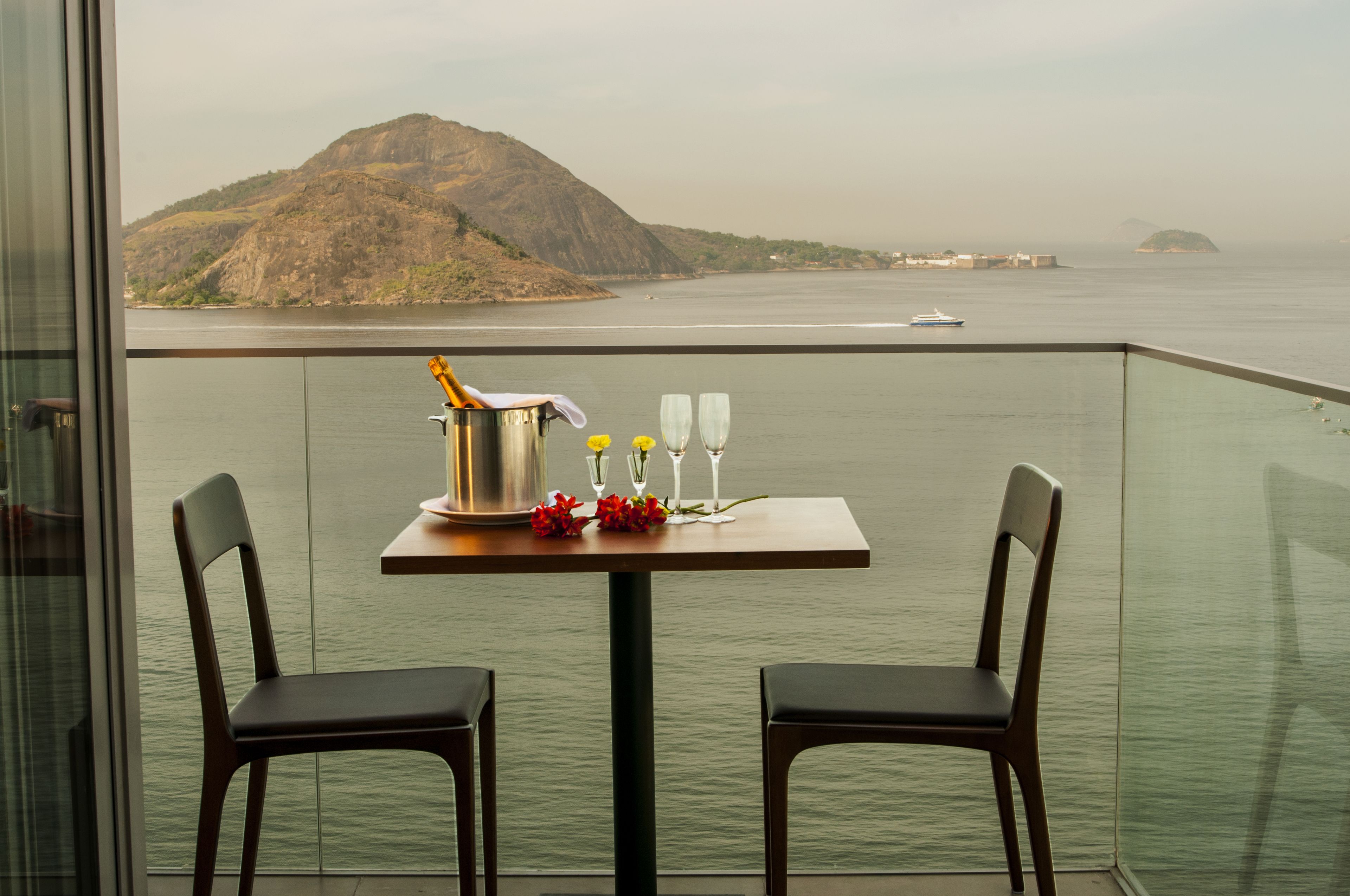 A balcony with a table set for two, overlooking a body of water and a distant mountain.