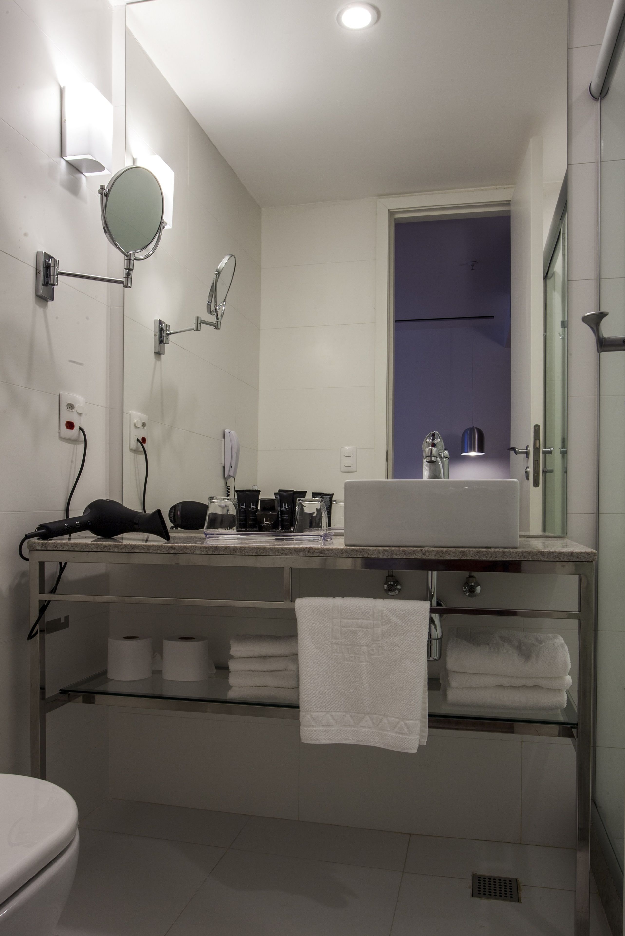 A modern bathroom with a white sink, mirror, and towel rack. There are toiletries on the counter and a shower visible through the door.