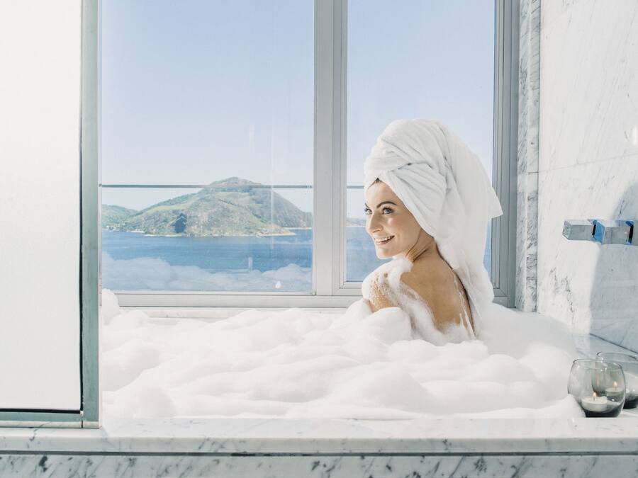 A person in a bathtub filled with bubbles, with a view of a mountainous landscape through a window.