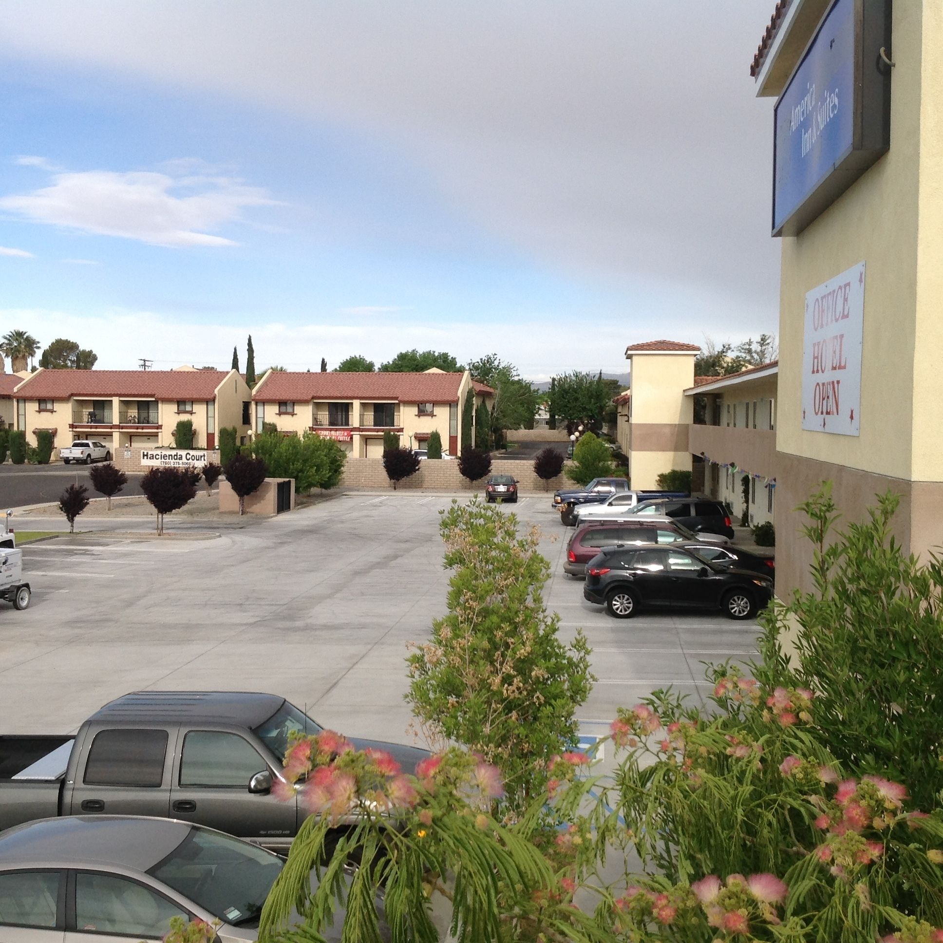 Um estacionamento com vários carros, um edifício com a inscrição "Hacienda Court" e um aviso indicando "Hotel de Escritórios Aberto".