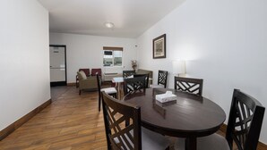 A dining area with a round table and six chairs, a window with blinds, a radiator, and a framed picture on the wall. - America Inn & Suites (Ridgecrest)