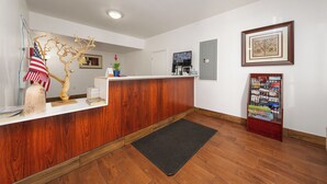 A reception area with a wooden reception desk, a black rug, a framed picture on the wall, and a bulletin board with various items. - America Inn & Suites (Ridgecrest)