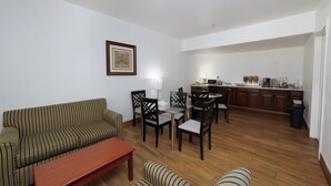 A living room with a sofa, chairs, a coffee table, and a dining area with a table and chairs. There is a kitchen in the background with cabinets and a countertop. - America Inn & Suites (Ridgecrest)