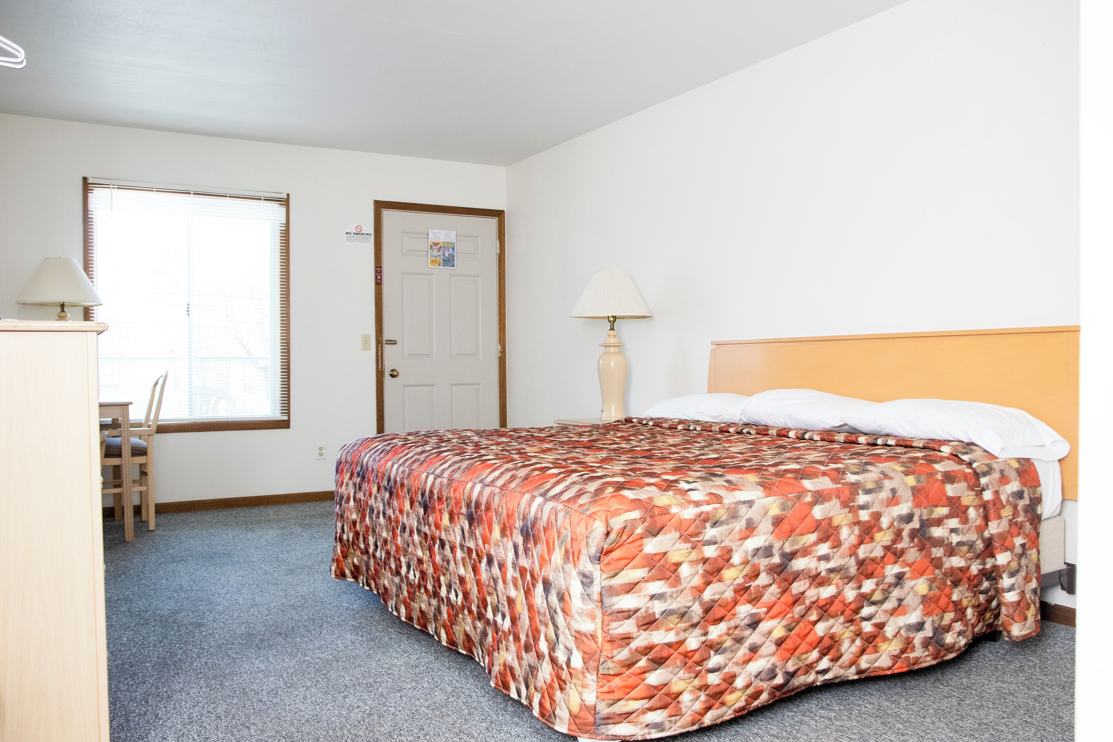A bedroom with a bed, a desk, a chair, a lamp, and a door.