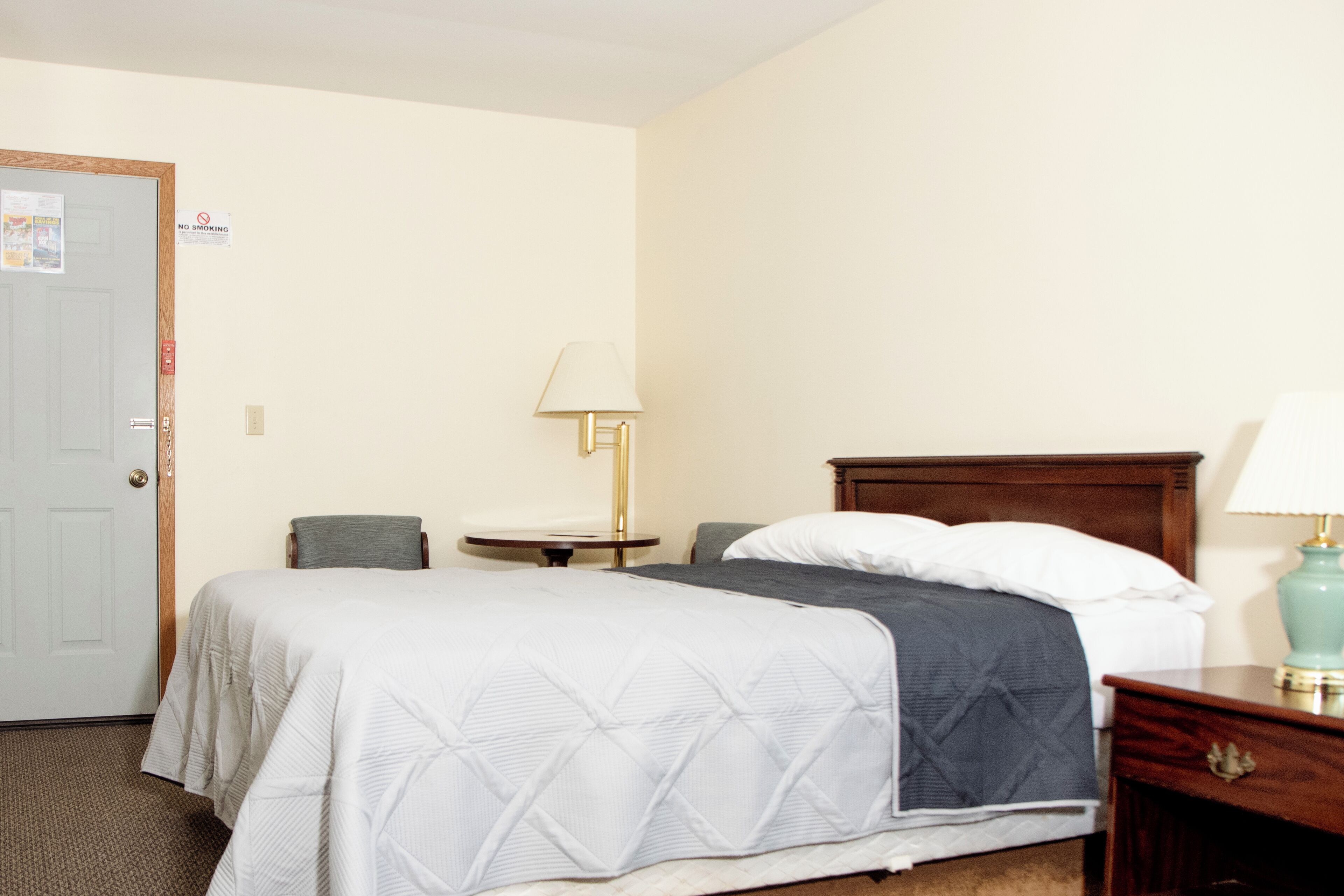 A hotel room with a bed, a bedside table, a lamp, and a door with a 'No Smoking' sign.