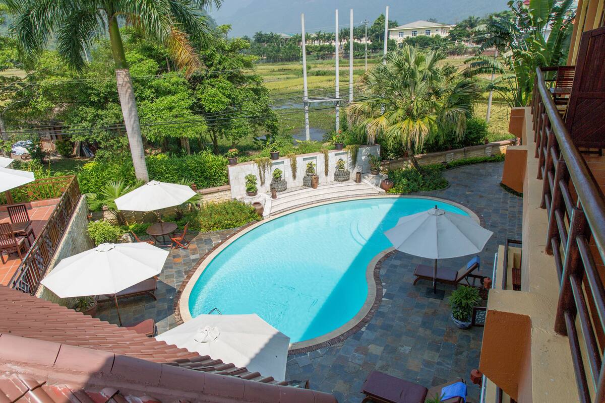 A swimming pool with lounge chairs and umbrellas, surrounded by lush greenery and a view of distant buildings.