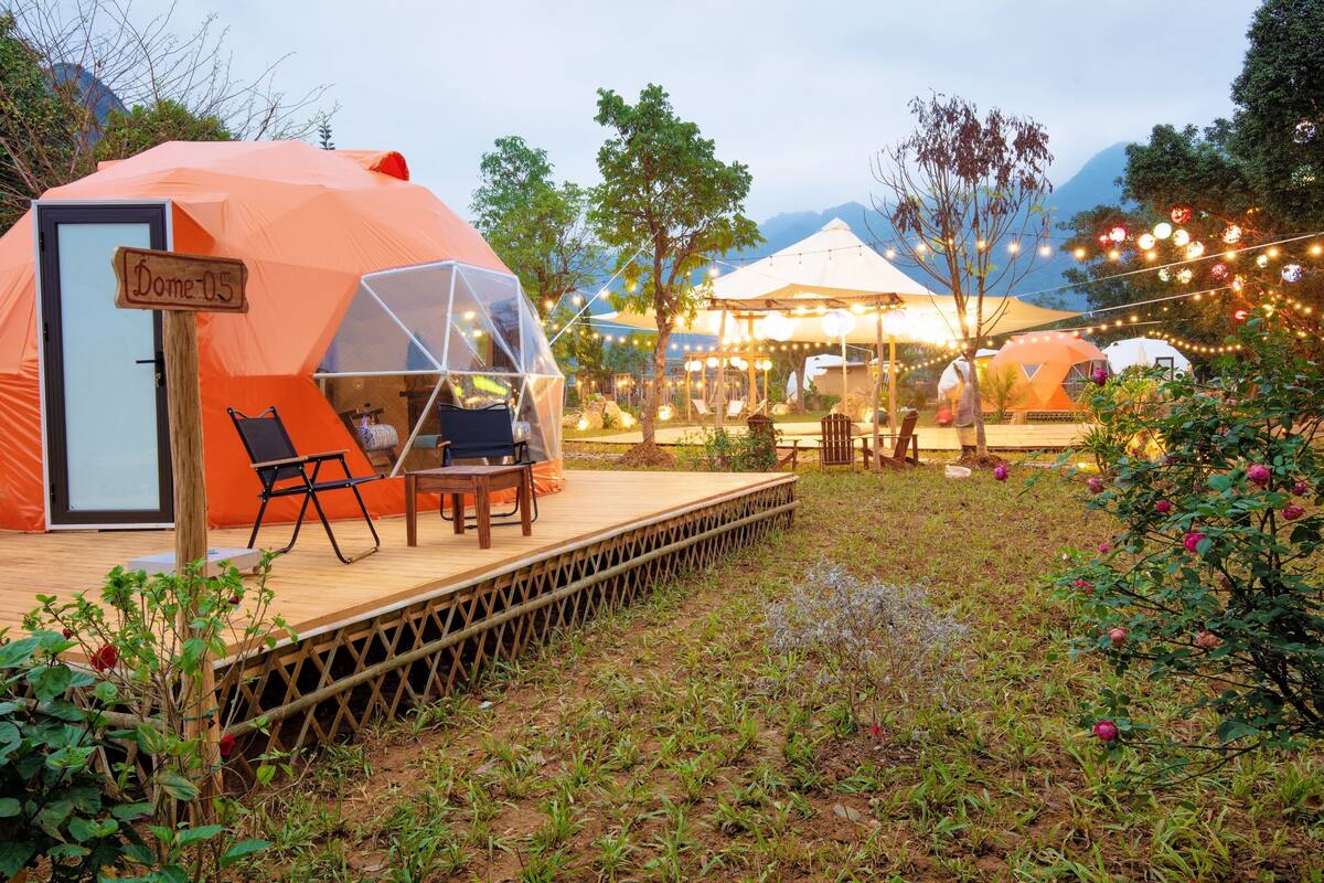 A dome-shaped tent with a wooden door labeled "Dome-05," a small deck with a table and chairs, and string lights in a garden setting.