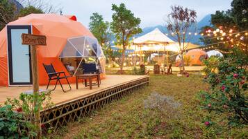 A dome-shaped tent with a wooden door labeled "Dome-05," a small deck with a table and chairs, and string lights in a garden setting.