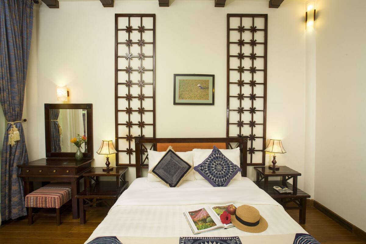 A hotel room with a large bed, two bedside tables with lamps, a wooden desk with a chair, a mirror, and a decorative screen.
