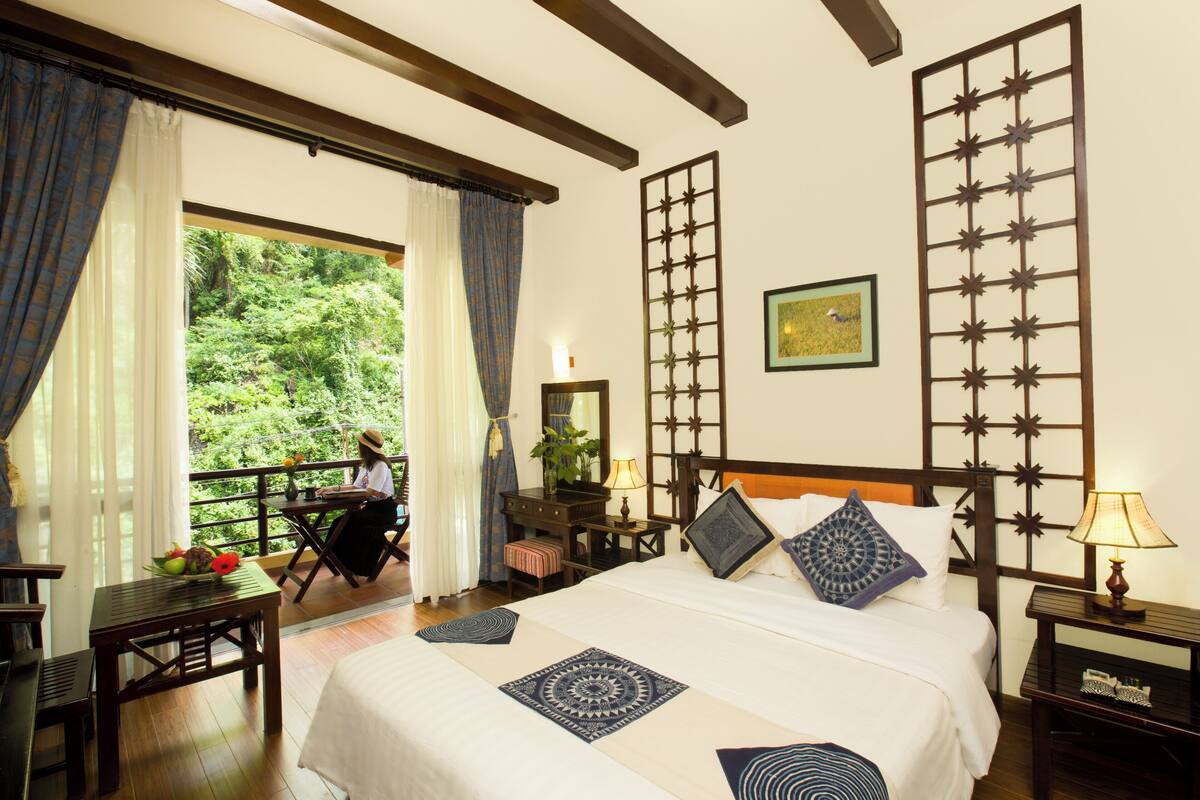 A hotel room with a large bed, a desk, and a view of lush greenery outside.