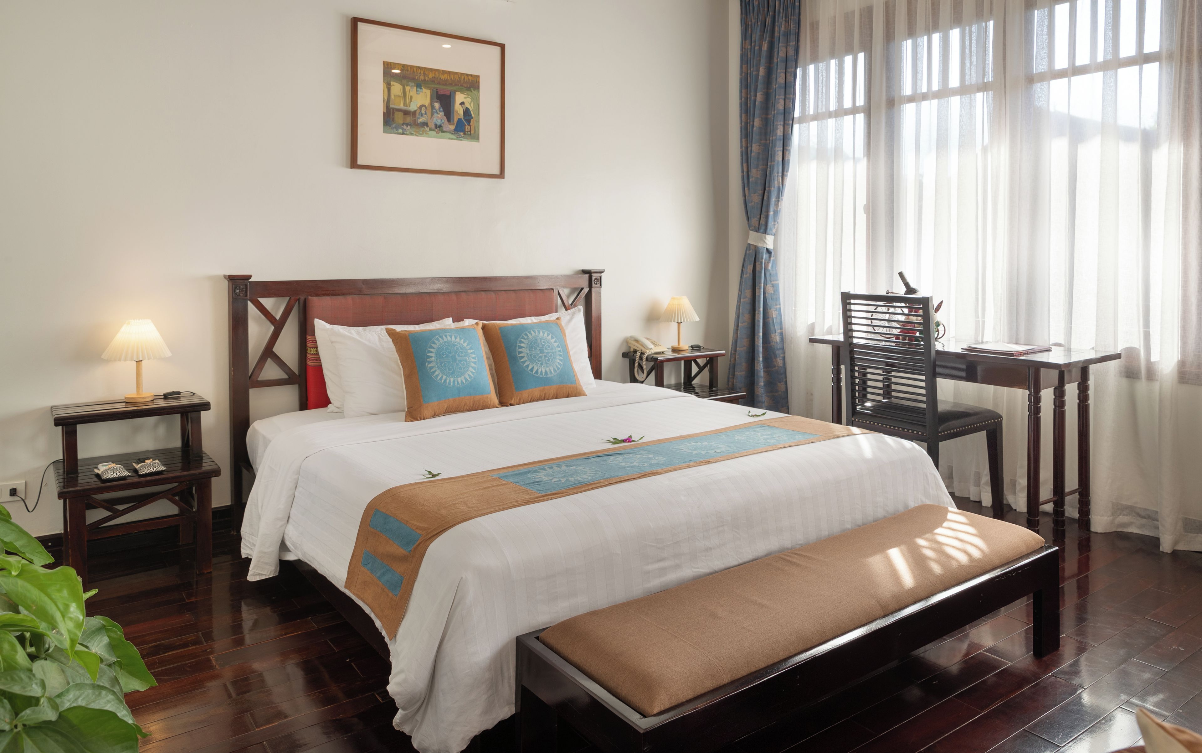 A neatly made bed with a wooden headboard, a bedside table with a lamp, a chair, and a desk with a lamp.