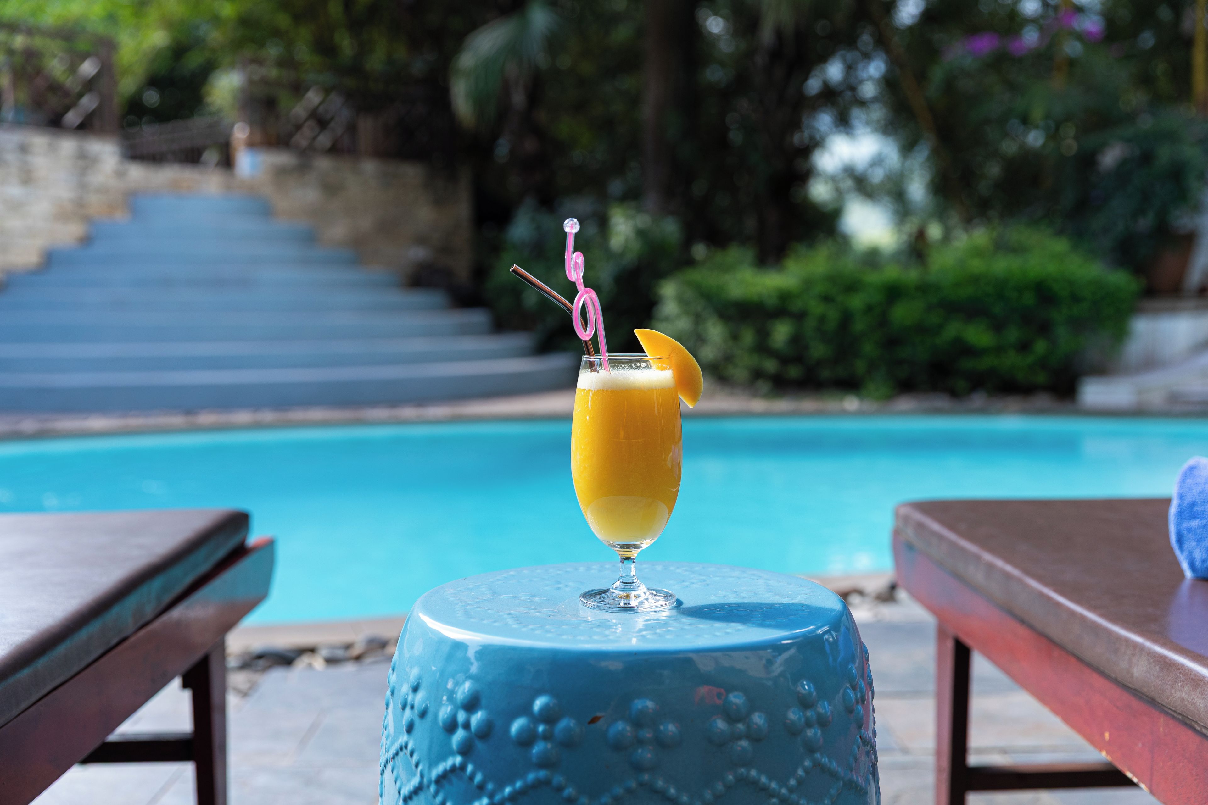Un vaso de jugo de naranja con una pajita y una rodaja de fruta sobre una mesa azul junto a una piscina.