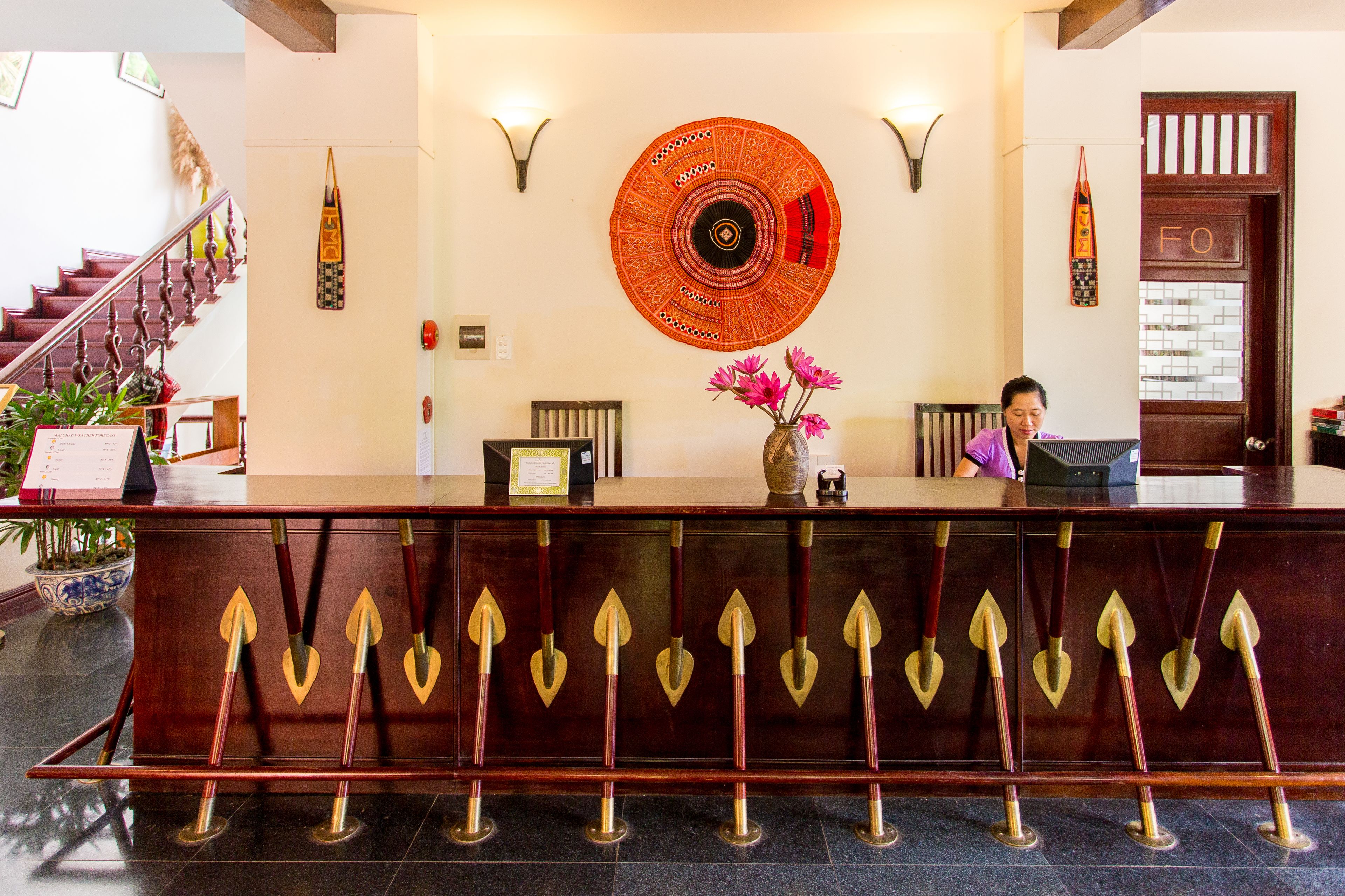 A reception desk with a wooden design, a large circular wall hanging, and a staircase in the background.