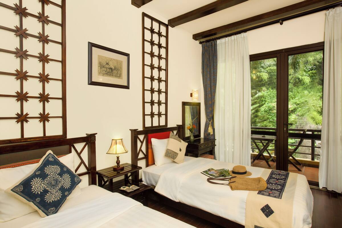 A hotel room with two beds, a wooden headboard, a nightstand with a lamp, a framed picture on the wall, and a window with a view of greenery.