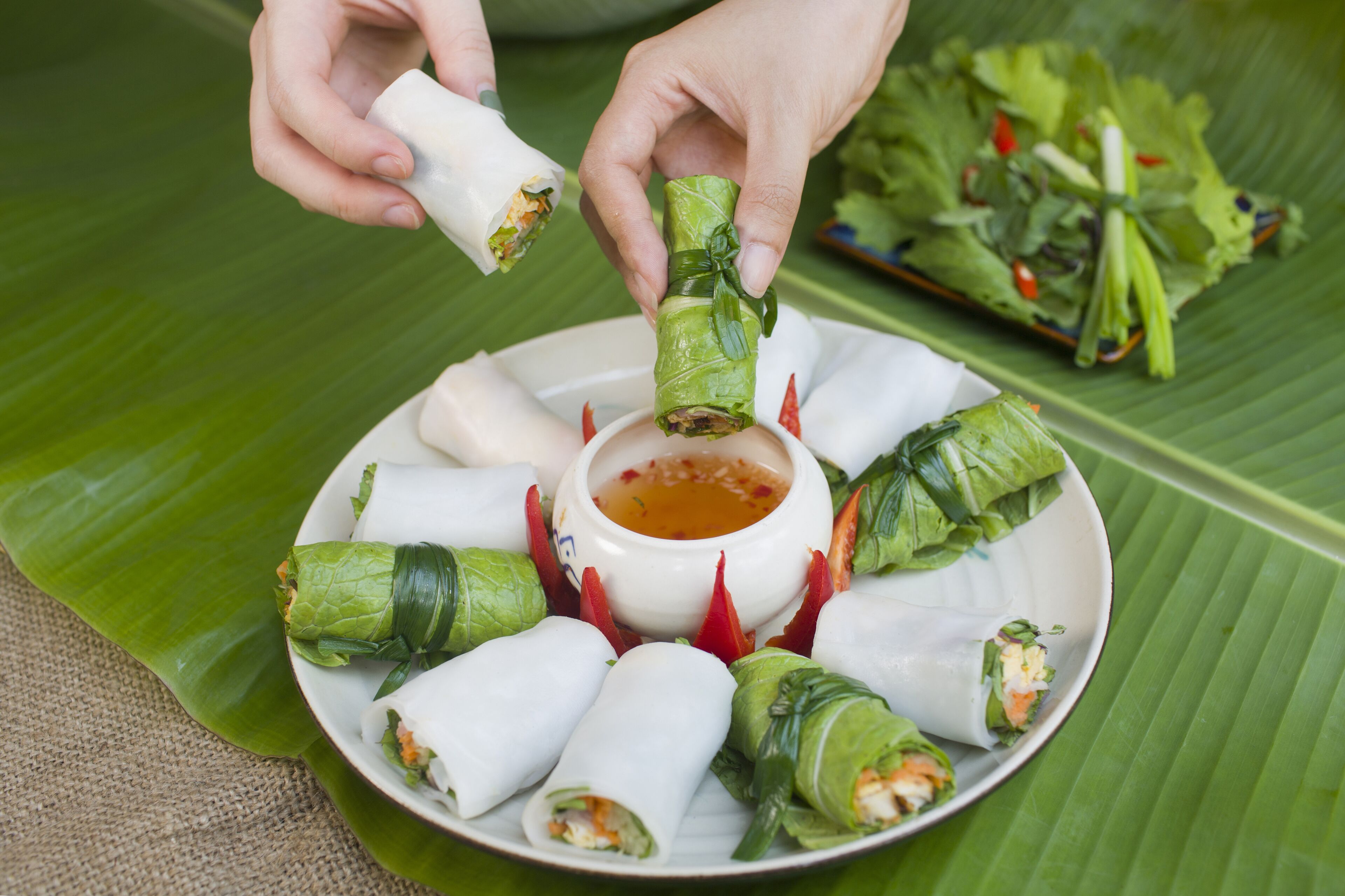 Une assiette de rouleaux de printemps accompagnés d’une sauce pour tremper, décorée de fines rondelles de piment rouge, le tout servi sur une feuille de bananier.