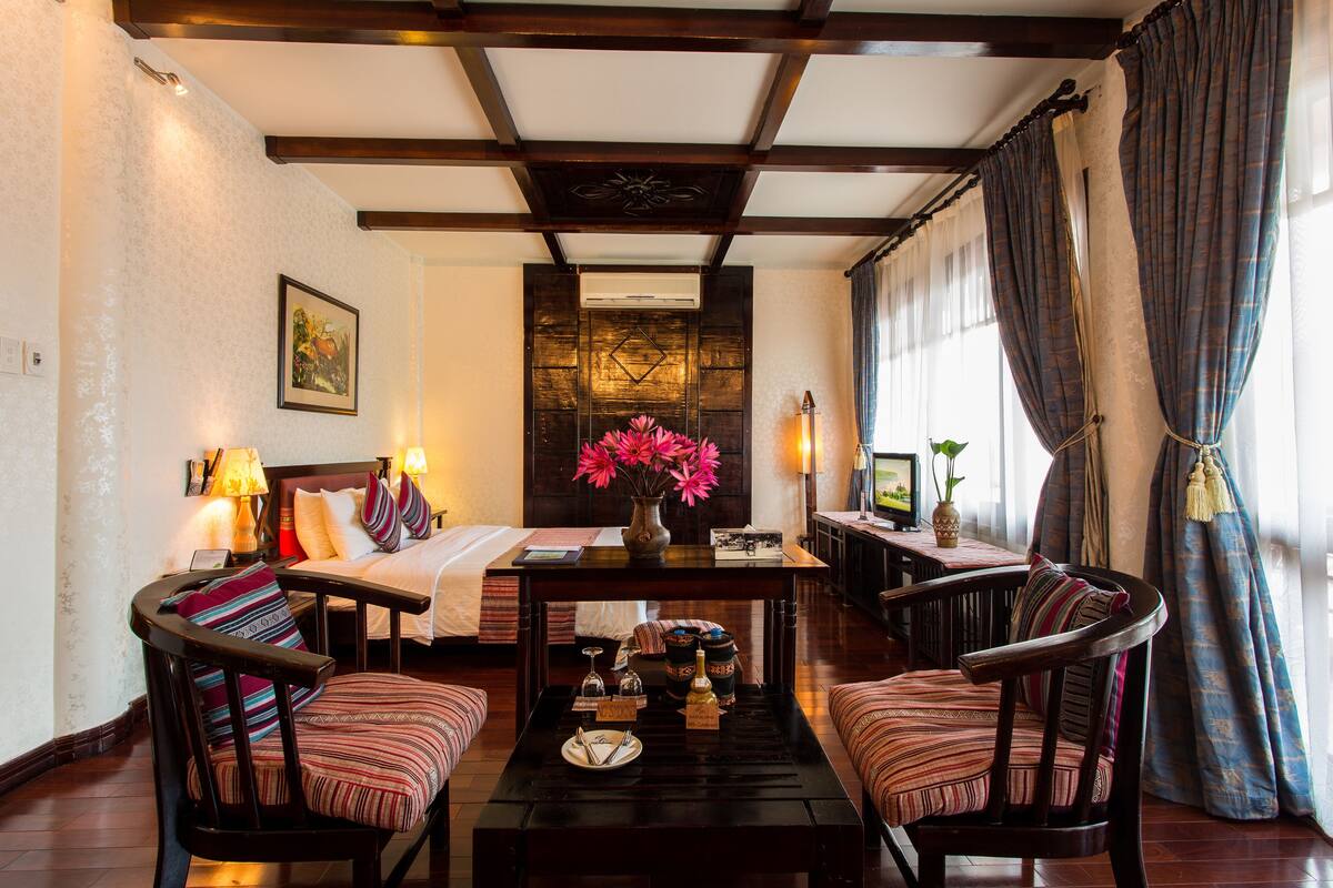 A cozy hotel room with a bed, two armchairs, a coffee table with decorative items, and a large window with curtains.