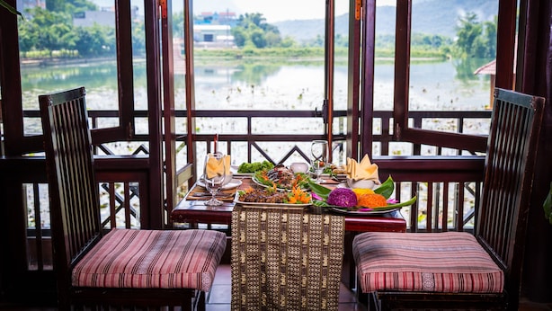 Un comedor con una mesa dispuesta para dos personas, con vista a un río y montañas y edificios a lo lejos.