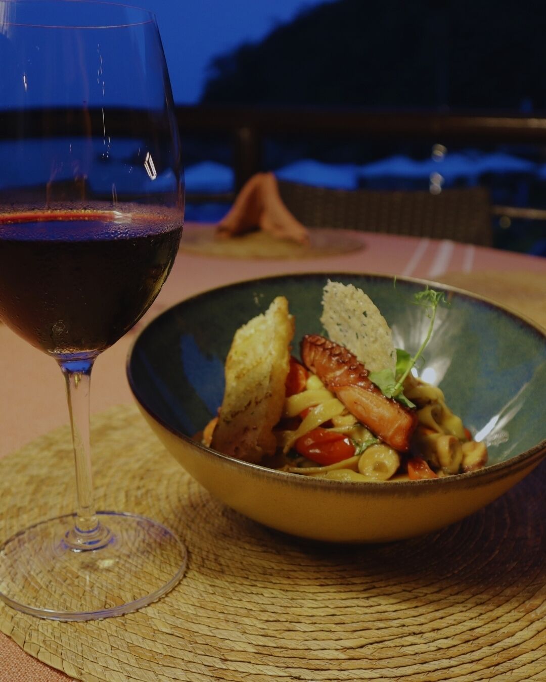 A bowl of pasta with vegetables and bread on a table with a glass of red wine.