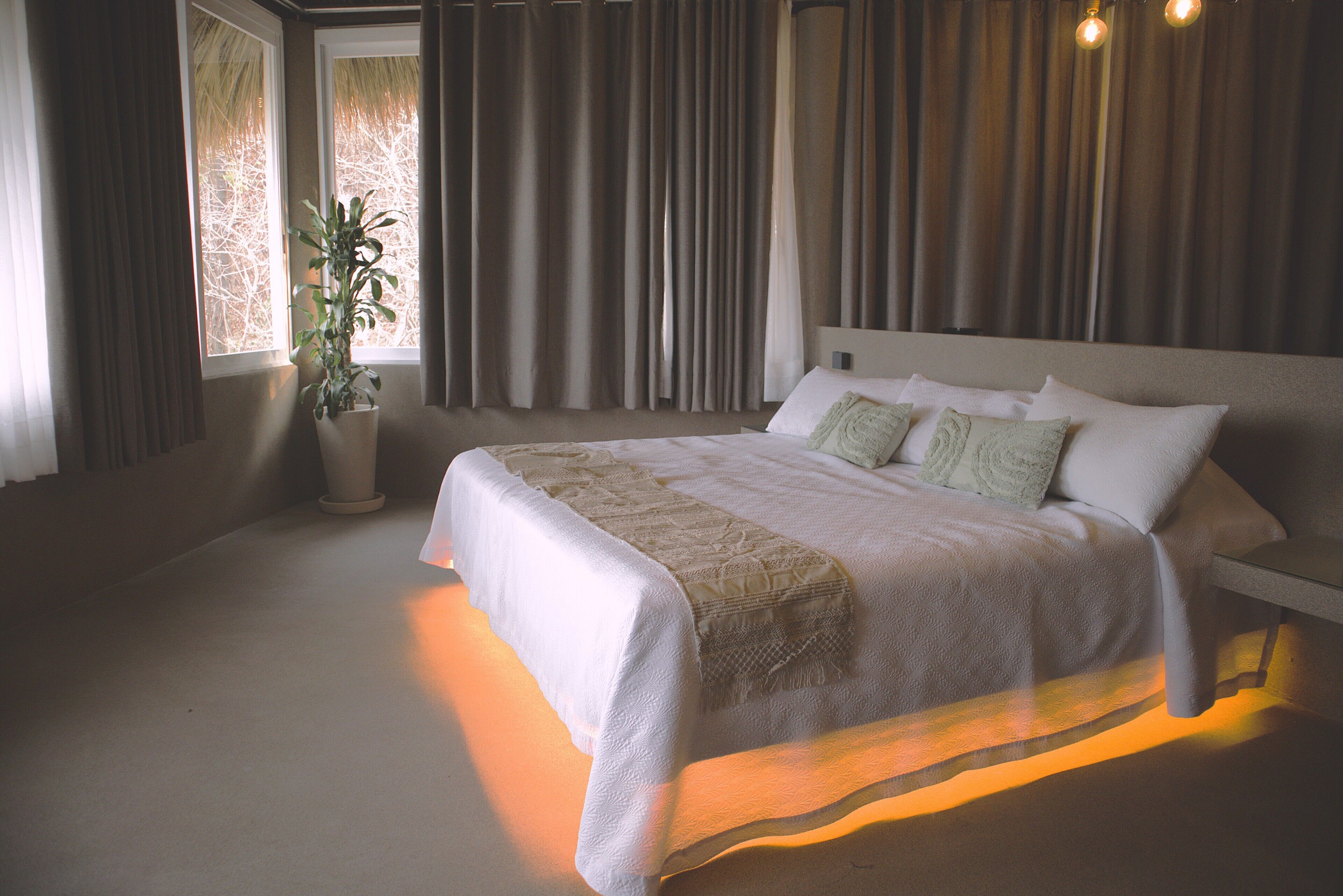 A neatly made bed with a white and beige bedspread, a potted plant, and a view of the outdoors through a window with curtains.