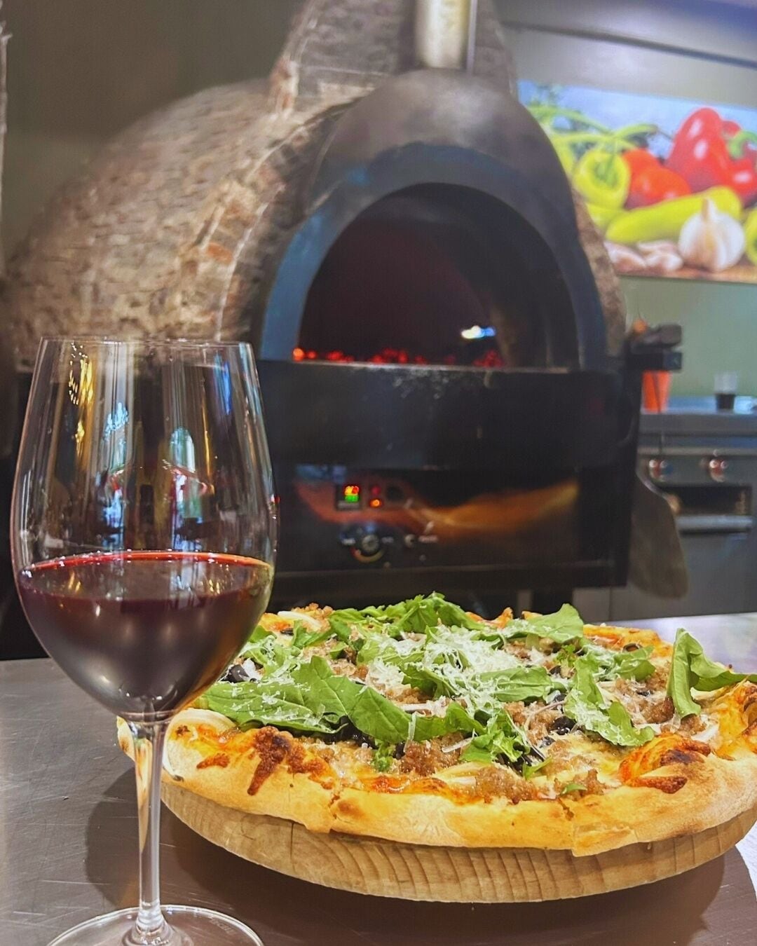 Una pizza con verdure e formaggio, un bicchiere di vino rosso e un forno a legna sullo sfondo.