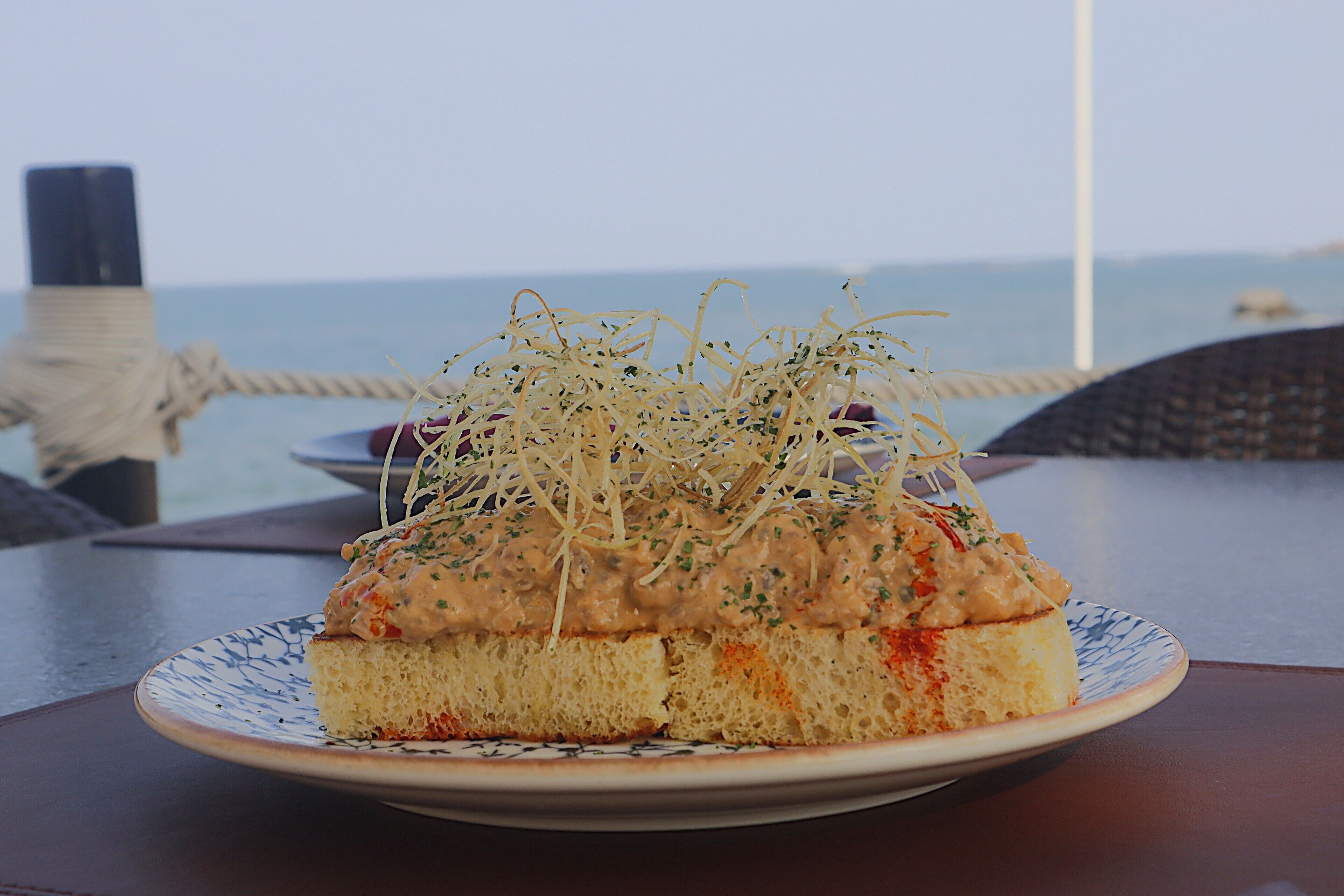 Een bord eten met een romige topping en geraspte garnering, op een tafel met uitzicht op zee.
